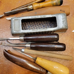 May include: A collection of woodworking tools, including chisels, a Pocket Surform, and a marking gauge. The tools have wooden handles and metal blades, arranged on a brown surface. The Pocket Surform is silver and has the text "STANLEY No. 398" and "POCKET SURFORM".