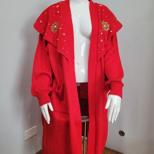 May include: A long, bright red cardigan sweater with a shawl collar. The collar is embellished with gold-colored sun and star designs. The sweater has long sleeves, pockets, and is displayed on a mannequin.