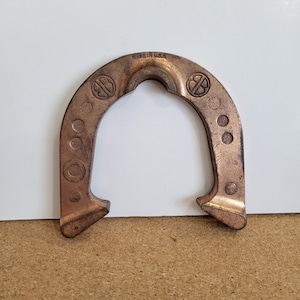 Peut inclure: Un fer à cheval de couleur cuivrée avec le texte "MADE IN U.S.A." est présenté sur un fond blanc et marron. Le fer à cheval a une forme incurvée avec des bords arrondis et plusieurs indentations circulaires. Deux logos circulaires sont visibles.