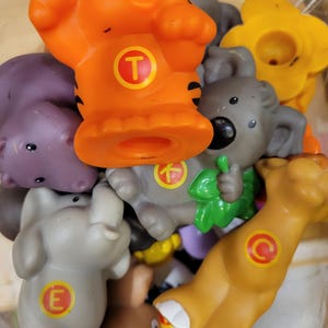 May include: A selection of brightly coloured plastic animal figures, including a tiger, koala, elephant, and lion. The tiger is orange with black stripes and a letter "T". The koala holds green leaves. The elephant has a letter "E".