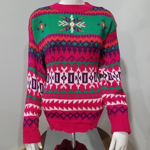 May include: A brightly colored sweater with a festive design. The sweater features a crew neck and long sleeves. The color scheme includes shades of pink, green, purple, and white, with geometric and snowflake-like patterns. The sweater is displayed on a mannequin.