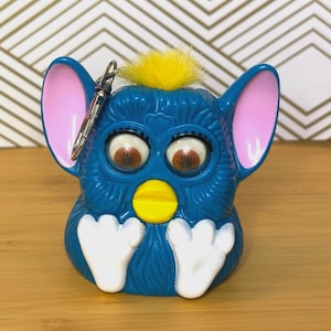 May include: A blue Furby keychain with pink ears, brown eyes, and a yellow beak. The Furby has white feet and a yellow tuft of hair on top. A silver keychain is attached to the top of the toy.