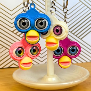 Peut inclure: Quatre porte-clés colorés inspirés de Furby en bleu, rose, blanc et violet. Chaque porte-clés présente un visage stylisé avec de grands yeux détaillés, un bec jaune et une bouche rose. Les porte-clés sont suspendus à des porte-clés argentés.