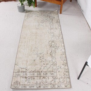 May include: A rectangular runner rug featuring a faded beige and cream base with intricate, distressed dark gray floral and geometric patterns throughout. The design includes a prominent border frame and a central medallion motif in a muted palette.