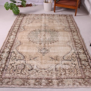 May include: A rectangular, distressed-look rug with a central medallion design. The rug features a beige background with intricate patterns in brown, green, and cream. The border is a darker brown with floral motifs, and the rug has a faded, vintage appearance.