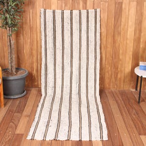 May include: A long, rectangular woven runner rug featuring a cream-colored base with repeating vertical dark brown stripes. The textile has a textured, slightly frayed appearance along the edges and a consistent striped pattern throughout its length.