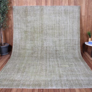 May include: A large rectangular area rug featuring a faded, distressed texture in shades of sage green and muted beige. The rug displays a subtle, worn-in pattern across its entire surface, with a low pile construction and finished edges.