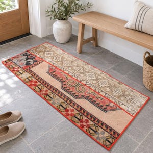 May include: A rectangular runner rug featuring a faded, multi-colored pattern with shades of red, beige, and black. The design includes geometric shapes, floral motifs, and a distinct border, laid flat on a gray tiled floor surface.