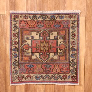 May include: A rectangular area rug featuring a central geometric medallion in red, tan, and dark blue. The rug has a wide border decorated with repeating floral motifs and a short white fringe along the top and bottom edges.