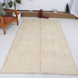 Puede incluir: Una alfombra beige claro con un patrón sutil y un borde rosa. La alfombra tiene un borde con flecos y está colocada sobre un suelo blanco.