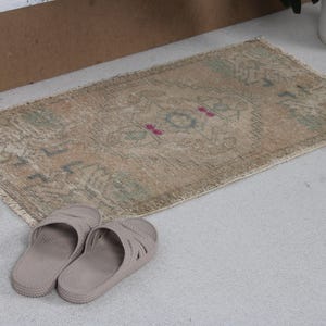 May include: A small, rectangular, distressed rug with a beige and faded color palette. The rug features a central design with pink and blue accents. Two pairs of light brown sandals are placed near the rug.