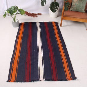 May include: A rectangular rug with a navy blue background and vertical stripes in orange, red, and white. The rug has a fringed edge and is placed on a white surface. The rug is a decorative home textile.