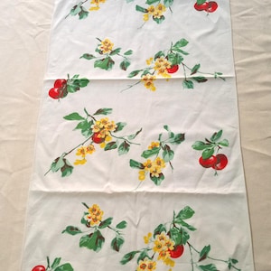 May include: A white rectangular table runner with a repeating pattern of red cherries, yellow flowers, and green leaves. The floral design is evenly spaced across the fabric, creating a vintage aesthetic. The runner is made of a soft, likely cotton, material.