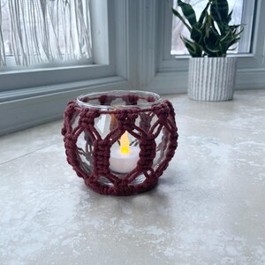 May include: A clear glass candle holder with a burgundy macrame design. The candle holder contains a white LED candle. The candle holder is sitting on a light-colored tile surface. A potted plant is visible in the background.