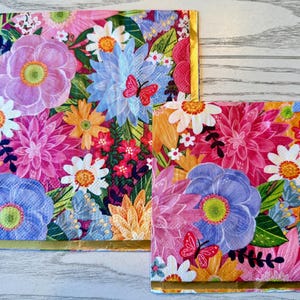 May include: Two floral napkins with a vibrant design. The napkins feature a colourful array of flowers in pink, purple, blue, orange, and white, with gold trim. Butterflies are also present in the design. The napkins are square and of different sizes.