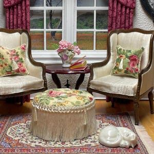 May include: Miniature living room scene with two cream-colored armchairs, floral pillows, and a round ottoman with fringe. A small table holds a vase of pink flowers and books. A decorative rug and a ceramic cat complete the scene.