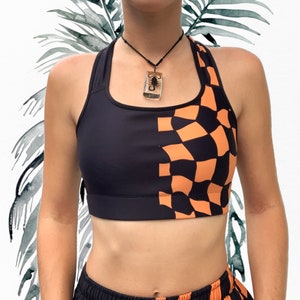 May include: Black and orange checkered sports bra with a halter neckline. The bra features a solid black panel on one side and a checkered pattern on the other. A black necklace with a scorpion pendant is also visible.