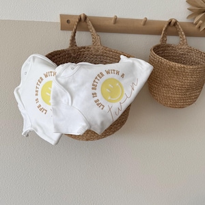Könnte beinhalten: Zwei weiße Baby-Bodys mit dem Text "Life is better with a twin" und einem gelben Smiley-Gesicht werden in einem geflochtenen Korb präsentiert. Der Korb hängt an einer Holzstange mit anderen dekorativen Gegenständen.