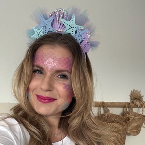 Könnte beinhalten: Eine Frau trägt ein weißes T-Shirt mit einem lila Muschel-Design und einem blauen Seepferdchen-Anhänger. Sie trägt ein Stirnband mit blauen und lila Seesternen und Muscheln. Ihr Gesicht ist mit einem Meerjungfrauen-Schuppen-Design bemalt.
