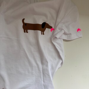 Könnte beinhalten: Weißes T-Shirt mit einer braunen Dackel-Illustration, schwarzem Ohr und einem rosa Croissant im Mund. Ein kleines rosa Croissant befindet sich auf dem Ärmel. Das T-Shirt besteht aus weißem Stoff.