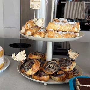 May include: A two-tiered white serving stand laden with pastries. The top tier displays cream-filled pastries, the bottom tier has cinnamon rolls. Wooden dove and church decorations are placed in the pastries.