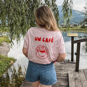 Könnte beinhalten: Hellrosa T-Shirt mit dem roten Schriftzug "UN CAFÉ" und einer Grafik eines Croissants und einer Kaffeetasse. Das Shirt wird mit Jeansshorts kombiniert. Die Person steht auf einem Holzsteg in der Nähe eines Sees.