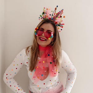 Könnte beinhalten: Ein festliches Outfit mit einem weißen Langarmshirt mit bunten Punkten, kombiniert mit hellblauen Jeans. Accessoires sind ein rosa Tüllschal, ein Haarreif mit Bommeln und passende Sonnenbrillen. Das Outfit ist verspielt und hell.