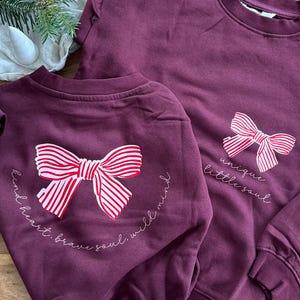 May include: Two matching burgundy sweatshirts. One has a large bow with red and white stripes and the words "kind heart, brave soul, wild mind." The other has a bow and the words "unique little soul."
