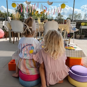Könnte beinhalten: Zwei Kinder sitzen draußen auf bunten Schaumstoff-Bodenkissen. Eines trägt ein weißes T-Shirt mit einer Grafik einer Gans mit Partyhut, Luftballons und dem Text "just two cool" über einem gestreiften Rock. Das andere trägt ein einfarbiges rosa Kleid.