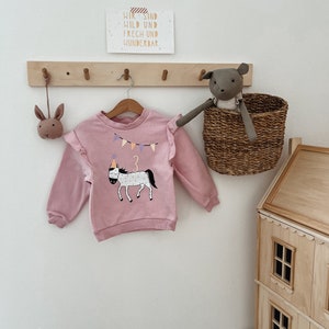 Könnte beinhalten: Hellrosa Kinder-Sweatshirt mit einem Pferde-Motiv, das eine Partyhut und die Zahl 3 trägt. Das Sweatshirt hat Rüschen an den Schultern und hängt an einer Holzgarderobe. Ein kleines Stofftier und ein Weidenkorb sind ebenfalls zu sehen.