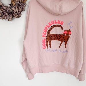 Puede incluir: Sudadera con capucha rosa claro con un diseño de leopardo de dibujos animados con gafas de sol y el texto "COOL PEOPLE CLUB FEEL FREE TO JOIN". La sudadera está colgada de una percha blanca.