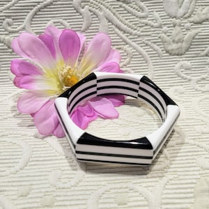 May include: A geometric, hexagonal bangle bracelet with a black and white striped design. The bracelet is set against a backdrop of a pink flower and a textured, off-white surface.