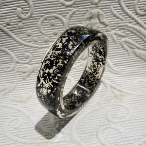 May include: A clear resin bangle bracelet with black and brown inclusions. The bracelet has a smooth, polished finish and a rounded rectangular shape. The inclusions create a unique, organic pattern throughout the bracelet. This accessory is suitable for everyday wear.