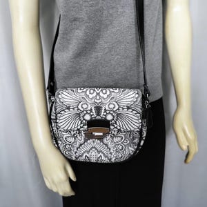 May include: A black and white crossbody bag with a decorative floral pattern. The bag has a black strap and a silver-coloured clasp. The bag is worn over the shoulder.