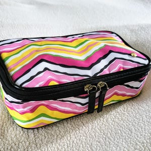 May include: A rectangular cosmetic bag featuring a colourful zigzag pattern in pink, yellow, white, and black. It has a black zip, black trim, and gold-coloured zip pulls. The bag is on a white, textured surface.