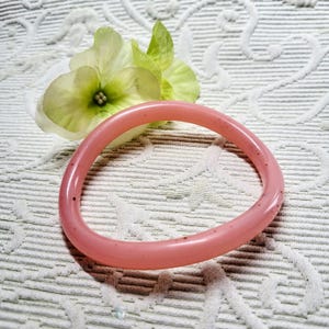 Op de afbeelding: Een lichtroze, ronde armband. De armband heeft een glad, glanzend oppervlak en ligt op een witte, getextureerde achtergrond. Een lichtgroene en witte bloem is ook zichtbaar.