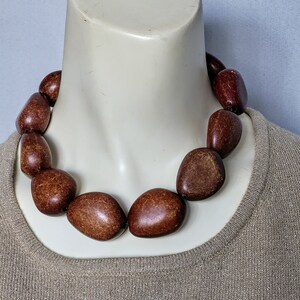 Puede incluir: Un collar llamativo con grandes cuentas ovaladas en tonos marrones. Las cuentas están ensartadas para formar una pieza audaz de joyería. El collar se exhibe en un maniquí, destacando su diseño y tamaño.