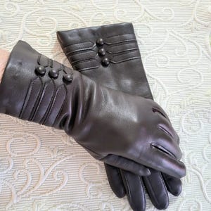 May include: A pair of dark brown leather gloves. The gloves have decorative stitching and three small, round buttons on the cuff. The gloves are designed with a classic, elegant style.
