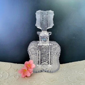 May include: A clear, cut-glass perfume bottle with a decorative stopper. The bottle has a square shape with intricate geometric patterns. A small cluster of pink flowers rests near the bottle.
