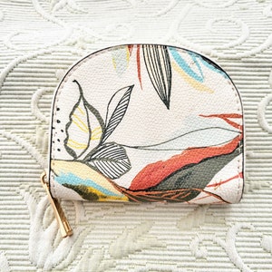 Könnte beinhalten: Eine kleine, halbkreisförmige Geldbörse mit einem farbenfrohen Blumendesign. Die Geldbörse hat einen cremefarbenen Hintergrund mit einem Muster aus Blättern und abstrakten Formen in Rot, Gelb, Blau und Schwarz. Sie hat einen goldfarbenen Reißverschluss.