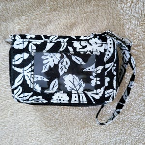 May include: A black and white quilted fabric wallet featuring a floral pattern and a clear plastic ID window on the front. The wallet includes a matching fabric wrist strap attached with a silver-toned metal clip and a zipper closure along the edge.
