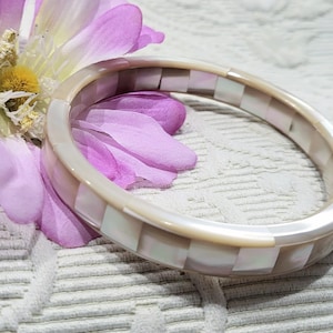 May include: A pearlescent bangle bracelet with a mosaic design, featuring alternating squares of white and light beige. The bracelet is resting on a white textured surface next to a purple and yellow flower.