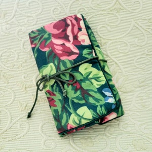 Op de afbeelding: Een opgerolde stoffen etui met bloemenpatroon, voornamelijk donkerblauwgroen, versierd met grote roze en bordeauxrode rozen, groene bladeren en blauwe accenten. Het is vastgebonden met een donkergroen koord. De etui is waarschijnlijk ontworpen om items op te bergen.