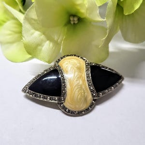 May include: A vintage brooch with a silver-tone metal frame. The brooch features a central cream-colored, swirled enamel design, flanked by black enamel triangles. The edges are lined with small, sparkling stones.