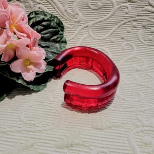 Op de afbeelding: Een levendige rode, gebogen armband gemaakt van een doorschijnend materiaal. De armband is een opvallend accessoire, met een gladde, glanzende afwerking. Roze bloemen en groene bladeren op de achtergrond voegen een vleugje natuur toe.