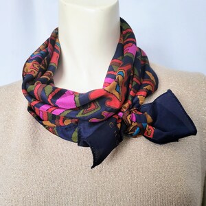 May include: A colorful silk scarf with a navy blue base and a vibrant pattern of pink, red, orange, green, and blue shapes. The scarf is draped around a mannequin's neck, with a knot and a label visible.