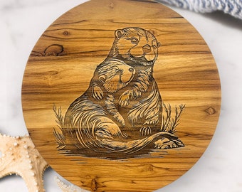 May include: Round wooden serving board with a laser-etched design of two otters embracing. The board has a natural wood grain pattern and is approximately 10 inches in diameter. The otters are depicted in a detailed, artistic style, with fine lines and shading.