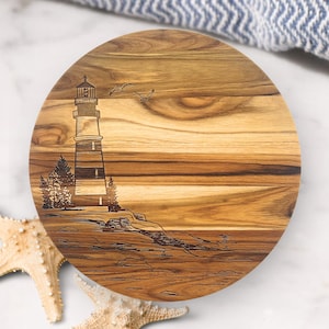 May include: Round wooden cutting board with a detailed engraved lighthouse scene. The lighthouse, with alternating dark and light bands, stands on a rocky cliff, with trees and seagulls. The board has a natural wood grain.