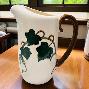 May include: A white ceramic pitcher with a dark brown handle and a spout. The pitcher is decorated with green ivy leaves and brown vines. The pitcher is sitting on a wooden surface.