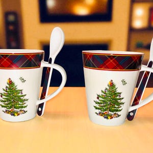 Könnte beinhalten: Zwei weiße Keramikbecher mit einem Weihnachtsbaumdesign und einem karierten Rand. Jeder Becher hat einen weißen Löffel, der auf dem Griff ruht. Die Becher stehen auf einer hellen Oberfläche.